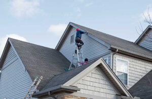 A homeowner clears leaves from a roof gutter beside a rain-soaked house.